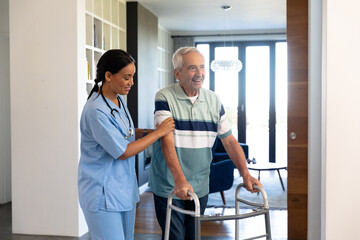 Biracial female physiotherapist assisting happy caucasian senior man in walking with walker at home - Powered by Adobe