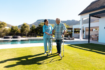 Biracial female doctor assisting caucasian senior man in walking with walker in yard on sunny day