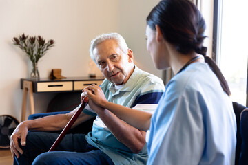 Biracial female physiotherapist consoling caucasian senior man sitting with walking cane on sofa