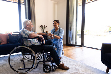 Biracial female physiotherapist talking and holding senior man's hand sitting on wheelchair at home - Powered by Adobe