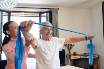 Biracial female physiotherapist assisting caucasian senior man stretching resistance band at home