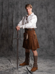 A young man in a leather kilt and a white lace-up blouse. A Scottish knight with a two-handed sword. Photo in the studio on a gray background