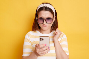 Horizontal shot of confused young woman wearing striped t-shirt standing with mobile phone and looking at display with puzzlement, posing isolated over yellow background.