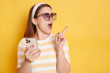 Image of excited amazed young woman wearing striped t-shirt standing with cell phone and pointing...