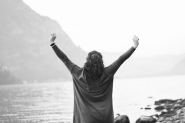 Black and white photo of happy girl standing on the beach and enjoying solitude on shores of Lake Lecco, Italy against the backdrop of mountains, water outdoors. Adventures. Blurred focus, rear view