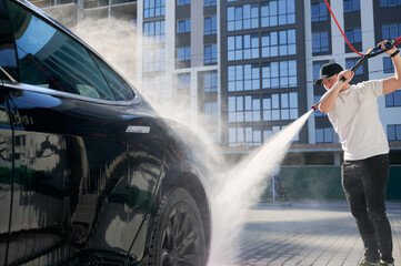 Man in cap cleaning his black modern car using high pressure water in service. Washing of auto with...