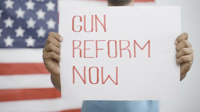 young man showing Gun reform Now sign board in front of US american flag - concept of protest against gun violence and activism.