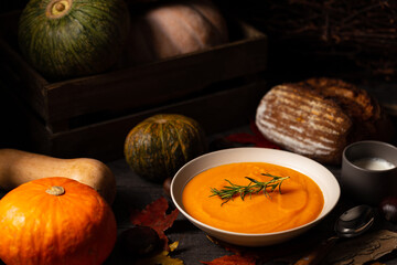 Pumpkin and chestnut soup with rosemary twig aside pumpkins, cream, bread and chestnuts on a vintage dark wood background spread with maple leaves.