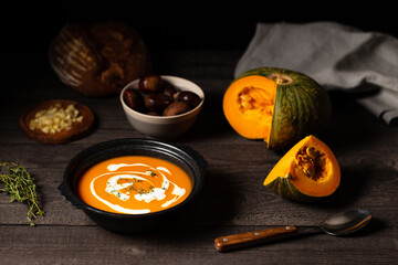 Pumpkin and chestnut soup with cream and thyme aside an open pumpkin, chestnuts and bread on a vintage dark wood background.