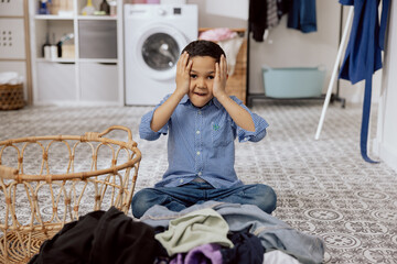 A child with dark hair sits on the laundry room floor, in the bathroom, a terrified boy clutching his head is doing housework, tiredly sorting through a huge amount of clothes.