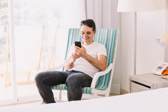 Young man sitting in a rocking chair in his bedroom with his legs crossed, looking at his cell phone, with light coming through the window. Concept of working, vacation, connection and smartphone.
