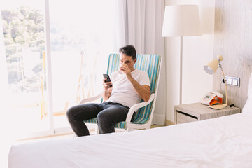 Young man sitting in a rocking chair in his bedroom, looking at his cell phone and drinking a...
