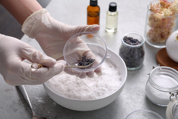 Woman in gloves filling bath bomb mold with dried flower buds at grey table, closeup
