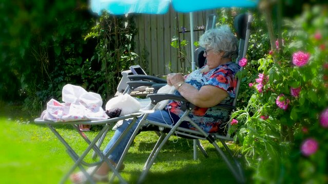 A senior woman &ndash; 85 years old &ndash; sitting knitting while relaxing in the shade on a lounger, in a peaceful, sunny garden / yard.