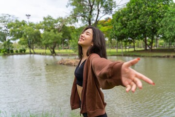 Young asian female lady model in her exercise sweater jacket sitting chillingly and calmly on the green grass while enjoying the calming breeze air and sunbath beside a calm lake in an outdoor park