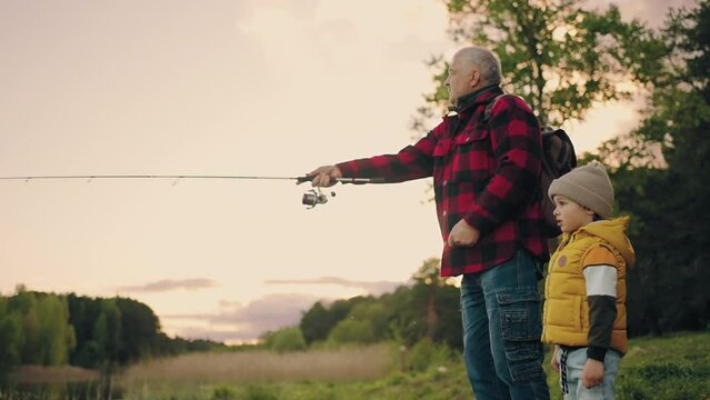 grandpa is teaching his grandson to fish on shore of lake, old man is throwing fishing rod