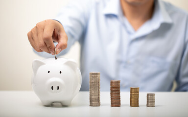 business man wearing a blue shirt putting coins into a white piggy bank, a pile of coins, finance,...