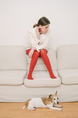 girl setting on sofa looking down. Sad pose mood.  beige and red. Small dog on the floor. excitement and anxiety sad emotions and experiences. 