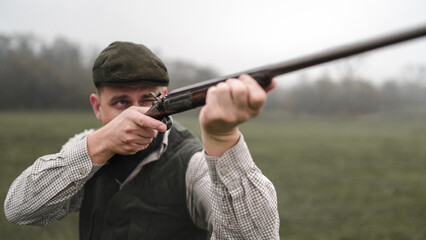 Hunter man in traditional shooting clothes on field aiming with shotgun.
