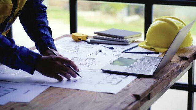 Young engineer looking at construction plans with blueprints Building or architectural drawings with rulers, pens, pencils, tape measure, architect's hats, and other desk accessories.