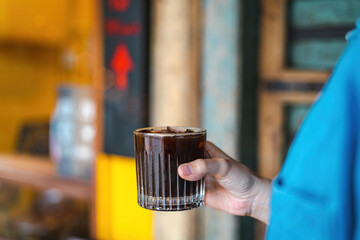 Hand holding a glass of ice coffee, americano in the afternoon over a vintage old wood door background at a Cafe coffee shop outdoor. Cold brew refreshment summer drink with copy space. Cafe concept
