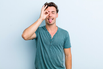 young handsome man smiling happily with funny face, joking and looking through peephole, spying on...