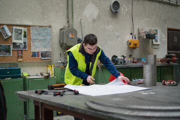 Fototapeta premium Young man with Down syndrome looking at blueprints when working in industrial factory, social integration concept.