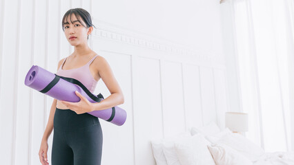 Fototapeta na wymiar Asian woman wearing sportswear and yoga pants carrying a rolled-up yoga mat standing in front of white wall background with sunlight from the window. Half-length image with copy space.