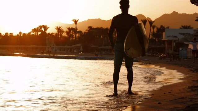 Silhouette man surfer in sunset light walking down beach with surfboard near waters edge enjoying summer time practice hobby during getaway vacations, guy recreating at tropical island with waves