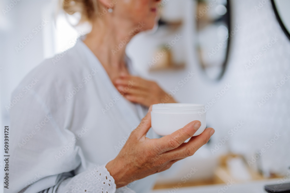 Wall mural Beautiful senior woman in bathrobe applying natural cream in bathroom, skin care and morning routine concept.