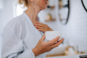 Beautiful senior woman in bathrobe applying natural cream in bathroom, skin care and morning routine concept.