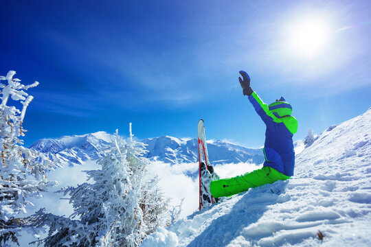 Boy with alpine ski sit in snow on Alps mountain top lift hands