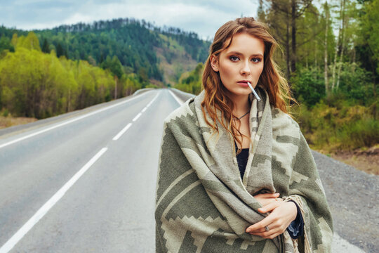 Portrait of a beautiful young girl wrapped in a blanket walking along the road with a smoking cigarette