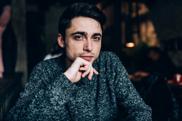 Half length portrait of handsome Caucasian millennial keeping hand at masculine beard during free time, good looking male dressed in casual clothes looking at camera and posing in coffee shop