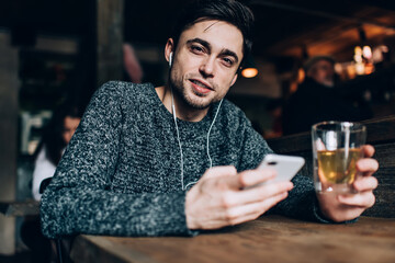 Portrait of young Caucaisan customer with beverage and smartphone in hands listening audio book during weekend daytime in cafe interior, millennial blogger in electronic headphones looking at camera
