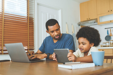 Black African American father working on laptop computer while his Afro Little son studying on digital tablet in kitchen at home.