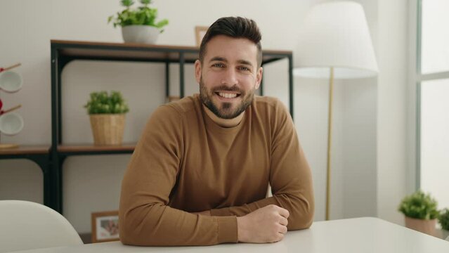 Young hispanic man doing ok sign with thumb up sitting on table at home