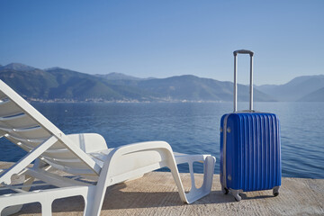 Blue suitcase next to a beach chair against sea and mountains, travel concept