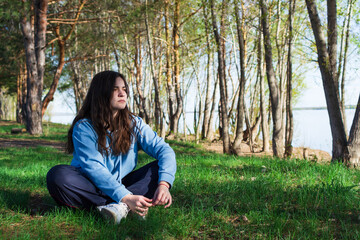 A young sad woman is sad, dressed in a blue hoodie, sits on green grass in the forest next to a pond, looks to the side and thinks about her life, copy space.