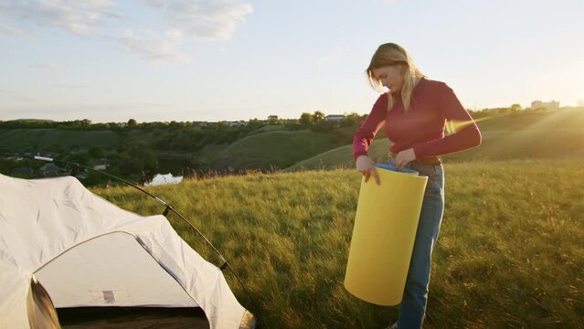 a young woman happily set up a tent on the mountain hills during sunset, for rest after a hike. a woman lays out a karimat. rest of one person in nature, in the mountains.