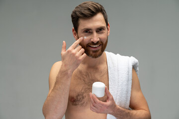 Bearded naked young man with perfect skin applying moisturiser facial cream isolated on grey...