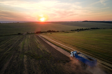 Aerial view of cargo truck driving on dirt road between agricultural wheat fields making lot of dust. Transportation of grain after being harvested by combine harvester during harvesting season