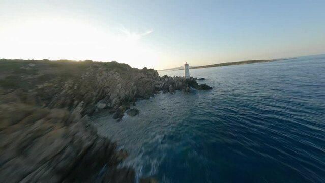 FPV video, view from above, stunning aerial view from an FPV drone flying at high speed over a rocky coastline with a lighthouse illuminated during a dramatic sunset. Faro di Capo Ferro, Sardinia.