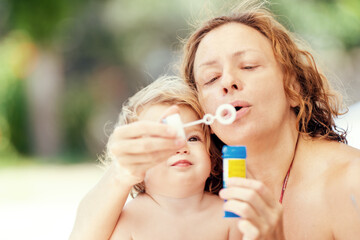 Fun summer day with family. Little girl plays with soap bubbles. Happy child blowing bubbles. Mom and daughter by the pool