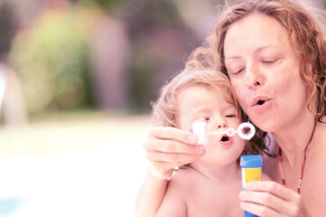 Fun summer day with family. Little girl plays with soap bubbles. Happy child blowing bubbles. Mom and daughter by the pool