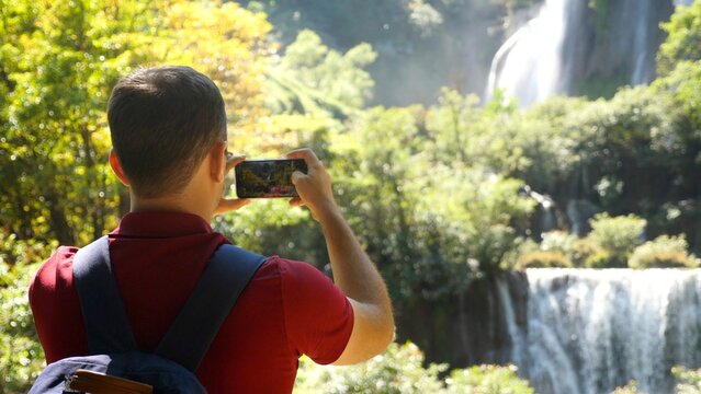 Young man traveler make photos for social media of amazing exotic waterfalls and tropical nature on his smartphone camera. Happy male tourist with backpack in travel. Wanderlust, tourism concept.