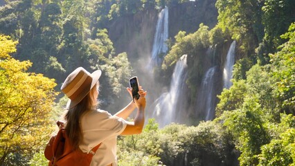 Young woman traveler make vertical video for social media of amazing waterfalls and tropical nature on her smartphone camera. Happy female tourist with backpack in travel. Wanderlust, tourism concept.