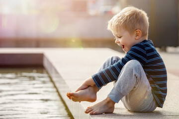 A curious child, a boy, barefoot touches the water of a city fountain. Hot and sunny summer day, the water is a good refreshment for the boy's feet.