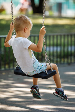 A Blond Boy In A White T-shirt Swings On A Swing In The Park On A Summer Day, Photo From Behind