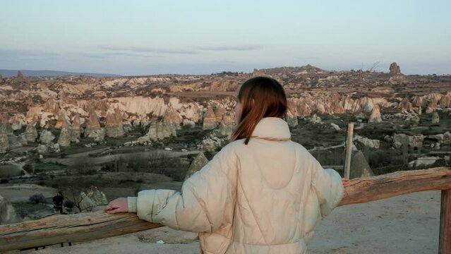 Back view of teenage girl standing on viewpoint, holding hands on wooden railing and enjoy stone valley scenery in old town Goreme Cappadocia Turkey on spring